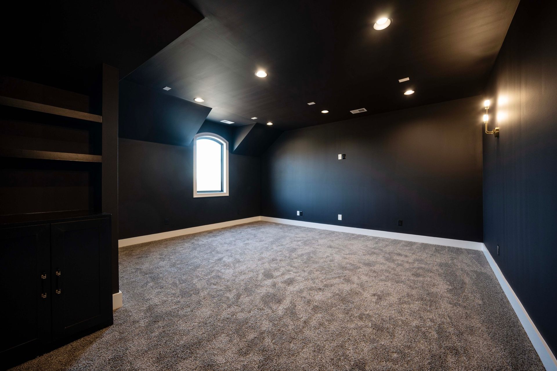 Black-walled room with gray carpet and small window. Built-in shelves and recessed lighting.