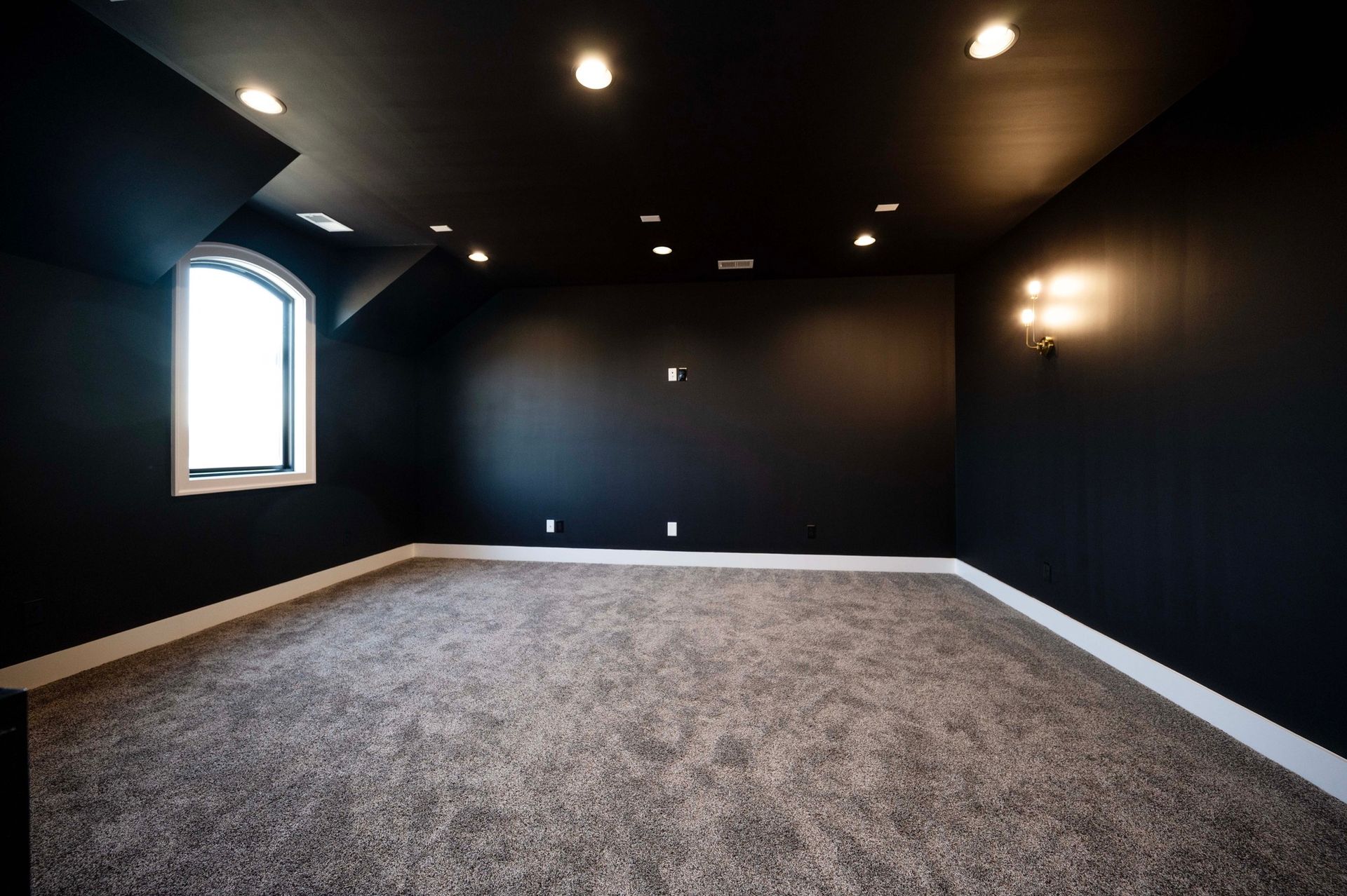 Black walled room, with a window and carpet flooring. Recessed ceiling lights.