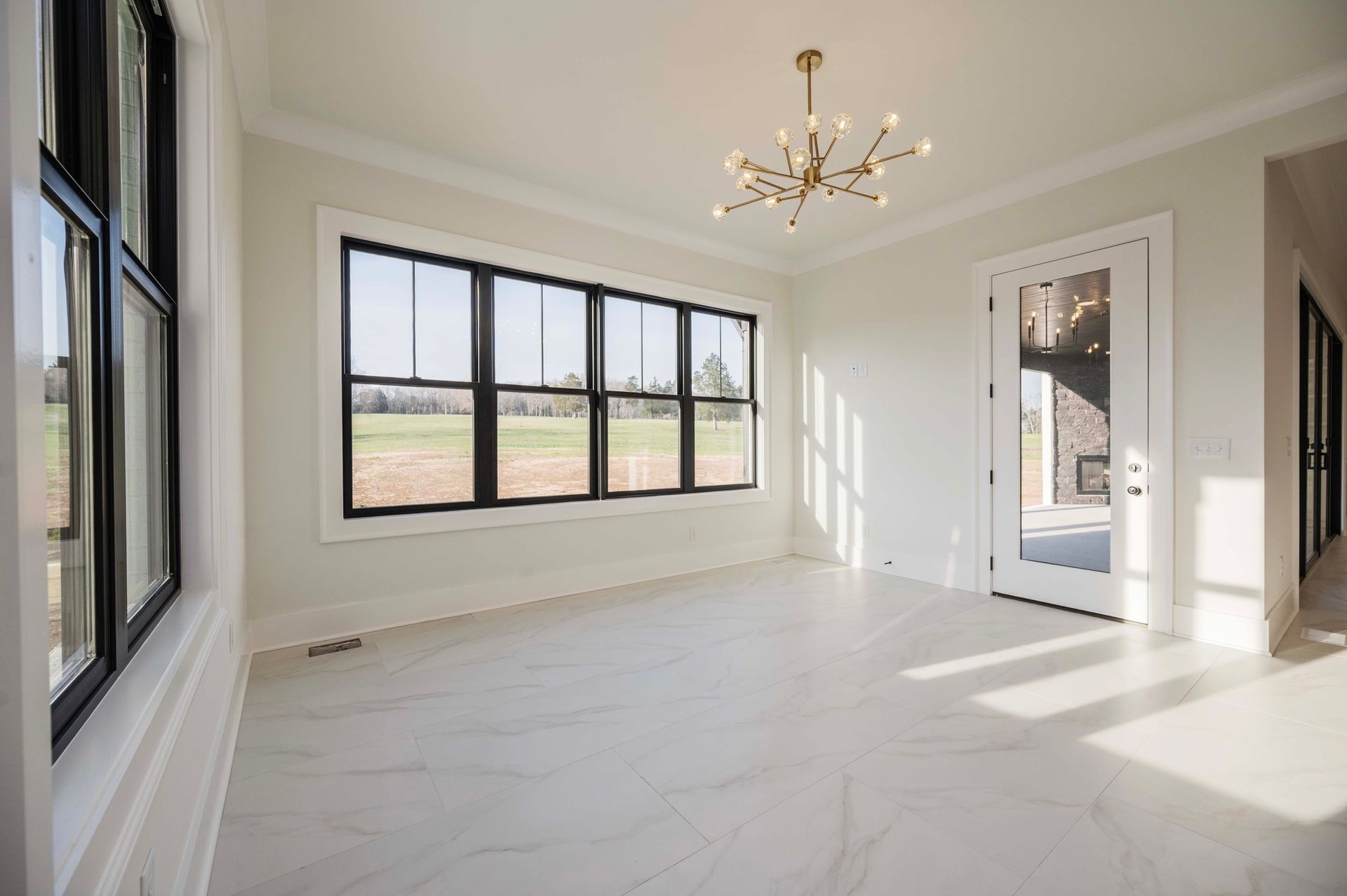 Empty room with large windows, black frames, and white tiled floor; chandelier hangs from ceiling.
