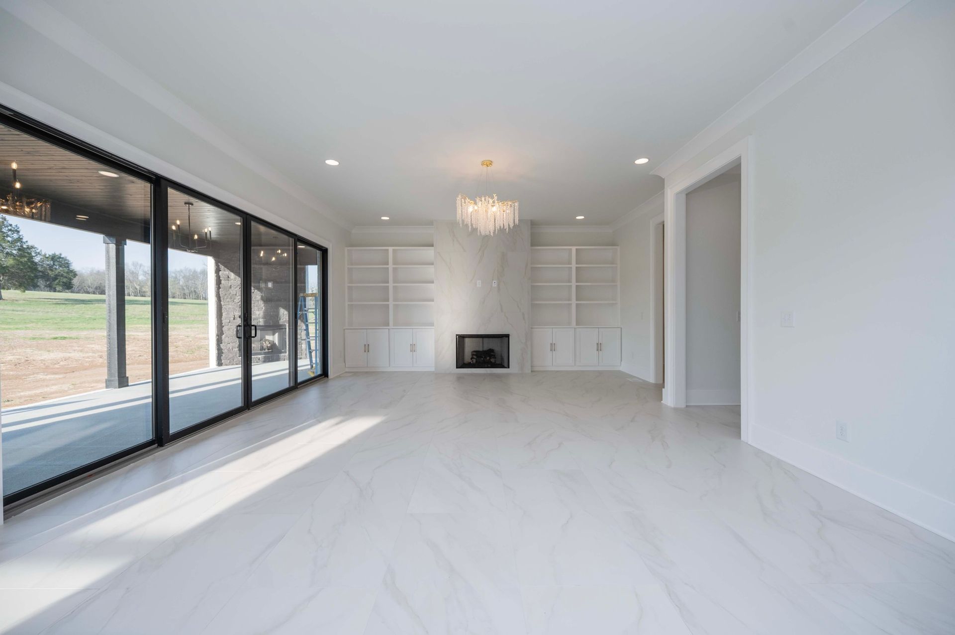 Empty, bright room with large glass doors. White marble floors, fireplace, and built-in shelving.