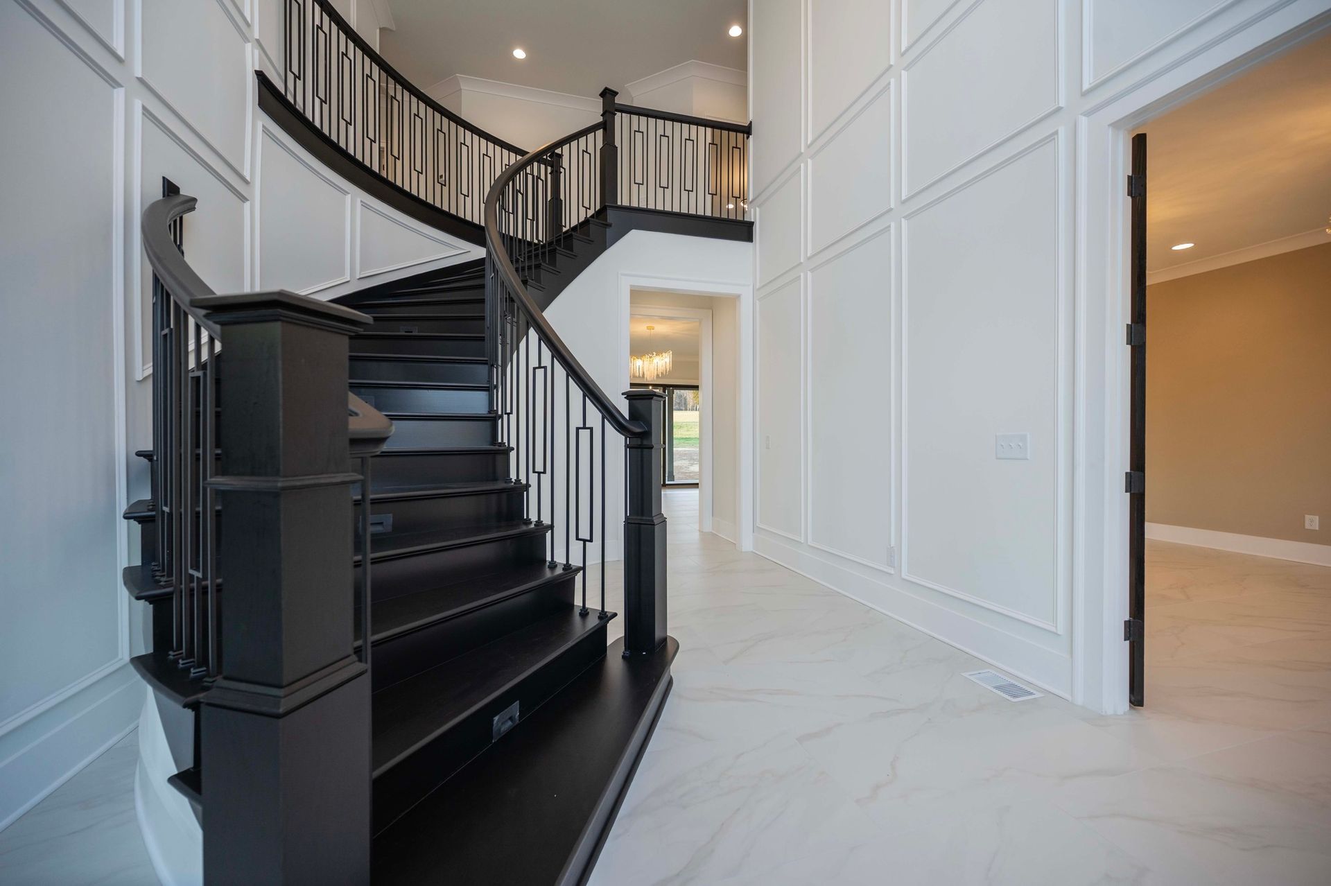 Black curved staircase in a bright white entryway with paneled walls and marble flooring.