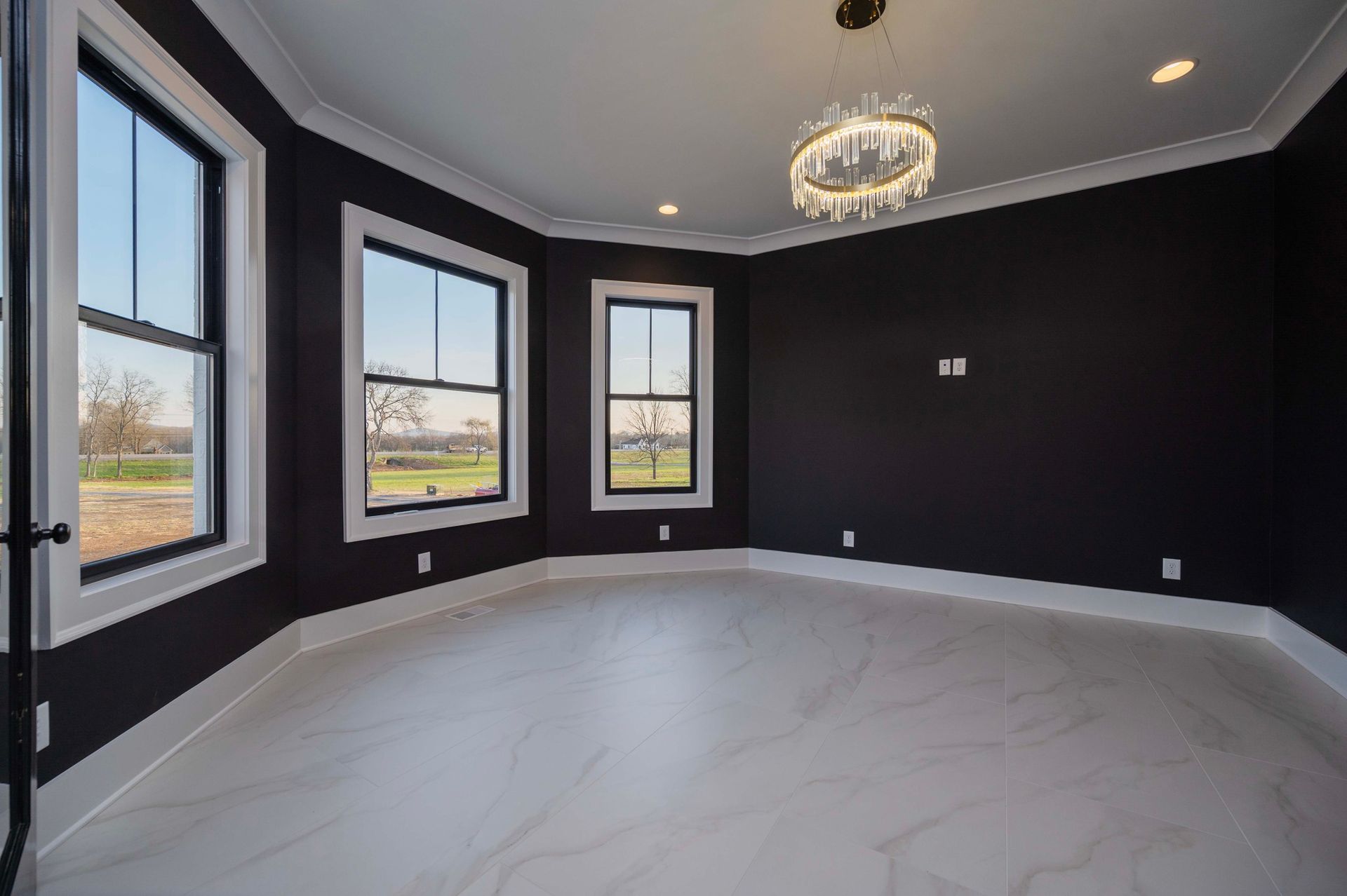 Empty room with dark walls, white trim, and light-colored flooring. Windows on left, chandelier.