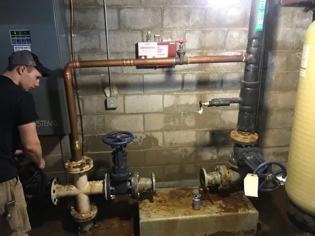 Man inspecting a basement boiler room with copper pipes, valves, and a water heater tank