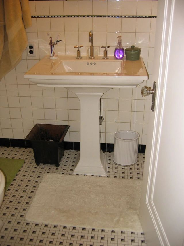 Small bathroom with pedestal sink, beige tiled walls, rug, and trash bin.