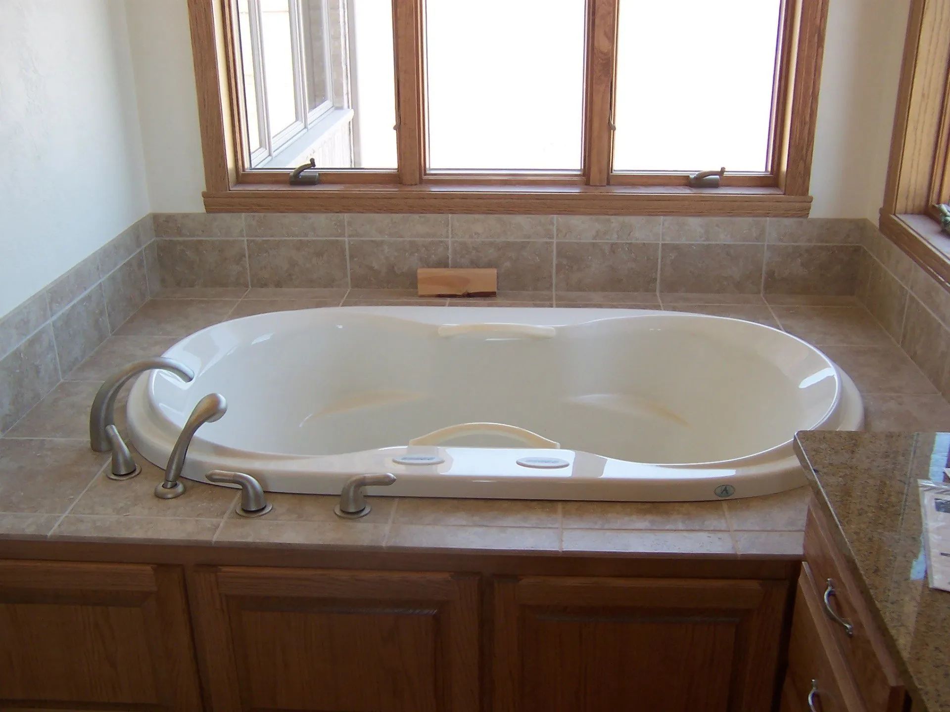 Built-in white jetted bathtub in a tiled bathroom with wood cabinets and a window above it