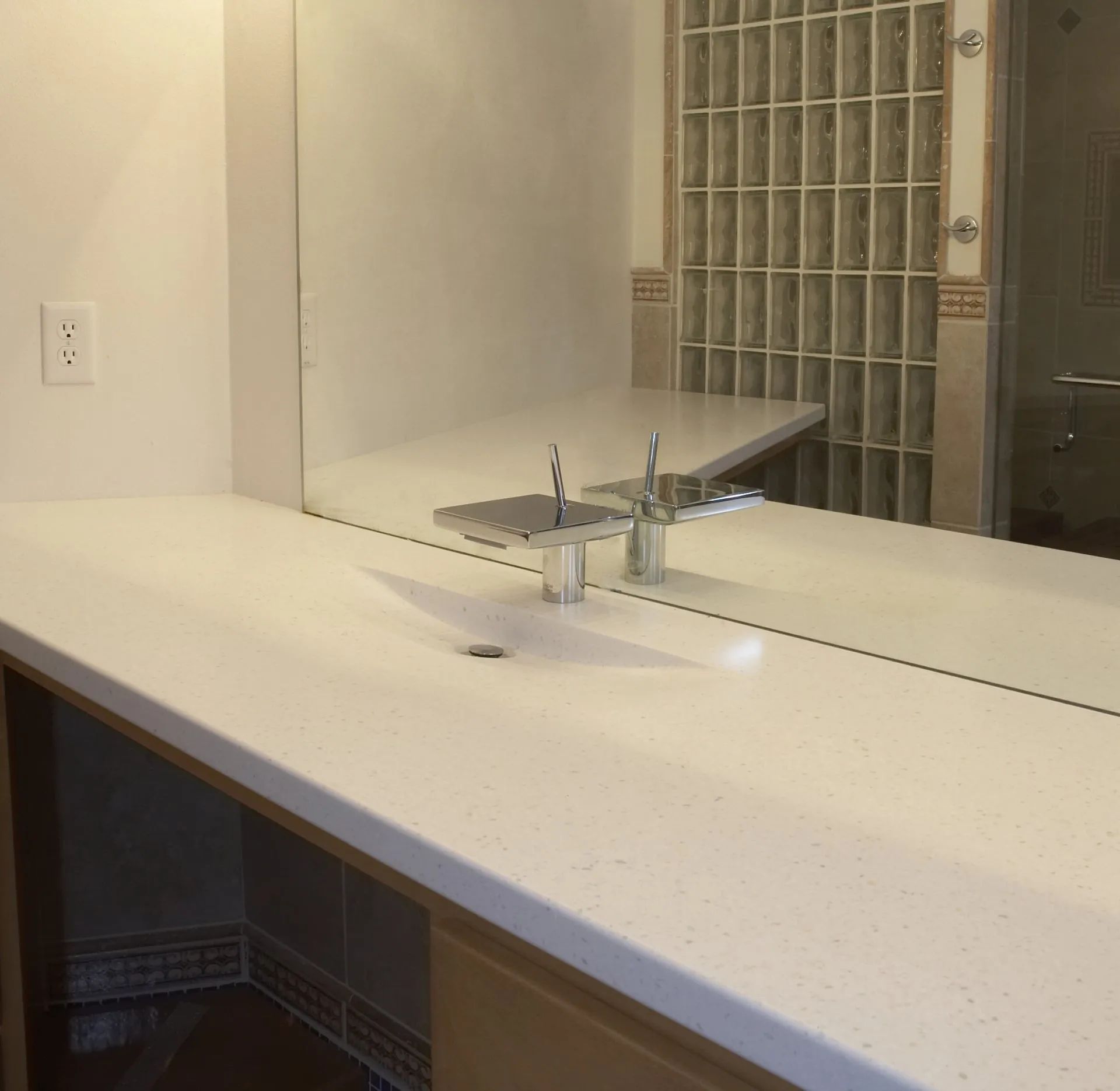 Bathroom vanity with white countertop, sink, and large mirror reflecting a glass-block shower wall.