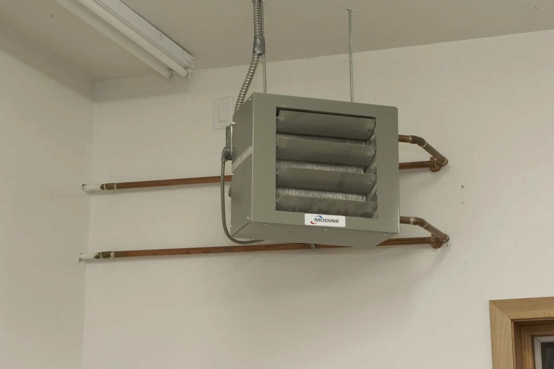 Wall-mounted gray heater/vent unit suspended by a chain in a plain room corner.