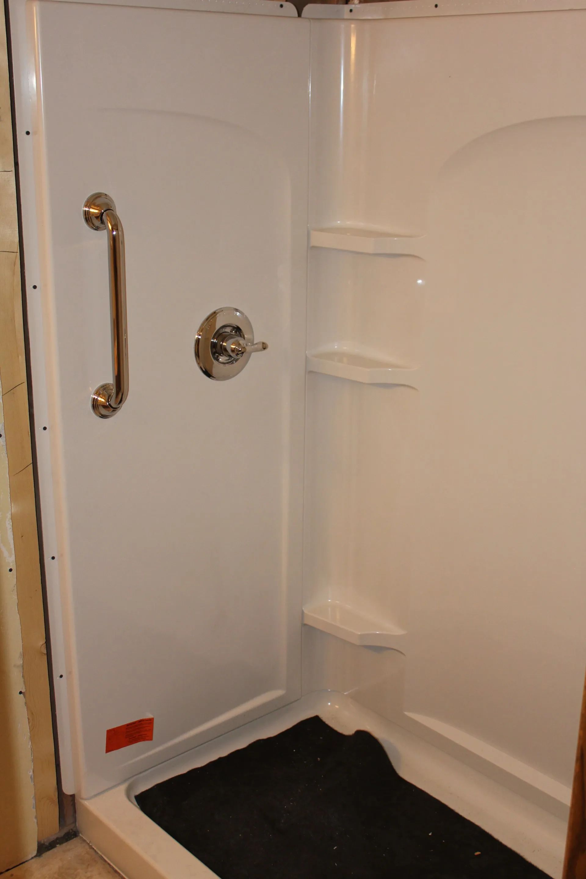 Corner shower stall with white walls, chrome grab bar, built-in shelves, and black floor mat.