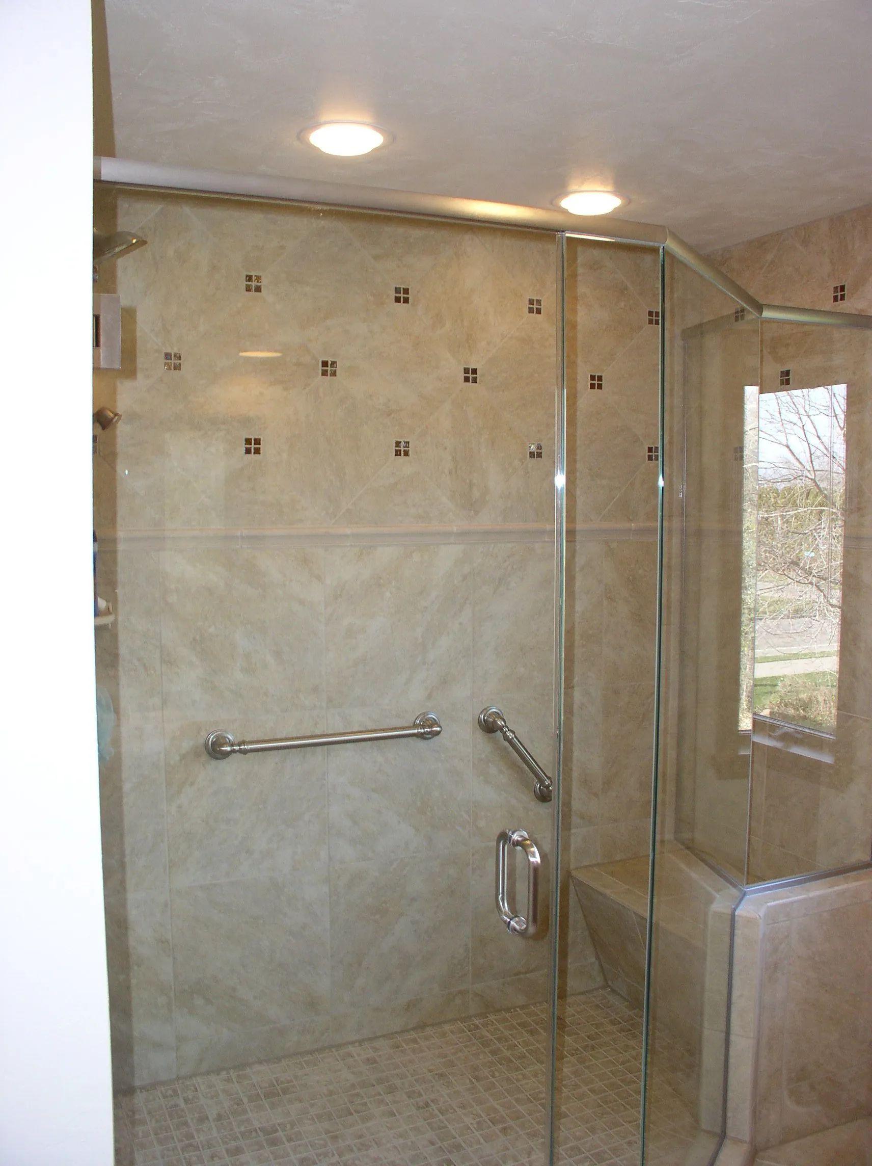 Glass-enclosed shower with beige tile walls, grab bar, and built-in bench under ceiling lights