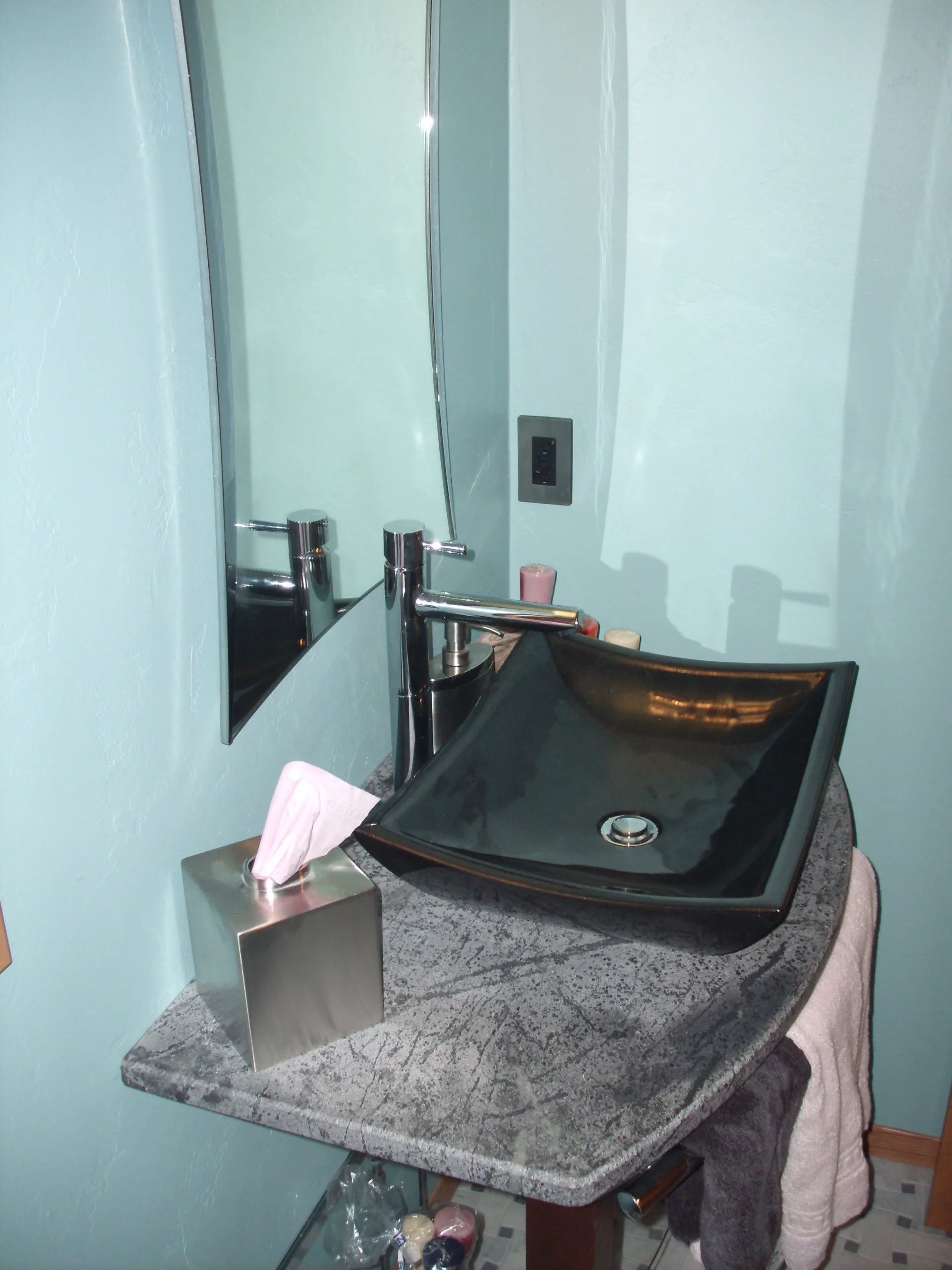 Bathroom sink and mirror with a dark basin on a gray countertop, next to a tissue holder.