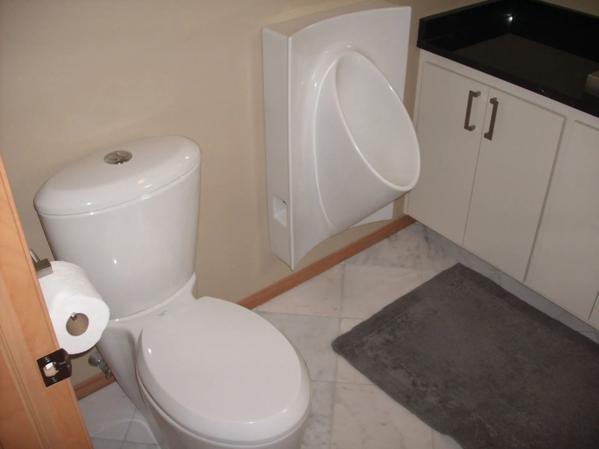 Small bathroom with white toilet, wall-mounted urinal, sink cabinet, and gray bath mat.