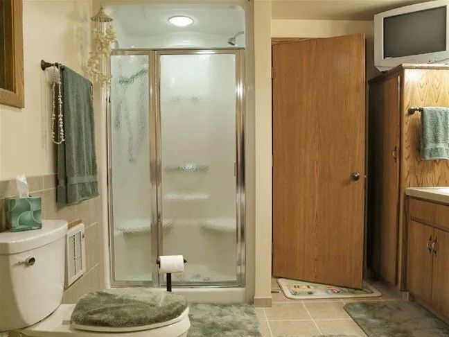 Small bathroom with shower, toilet, sink, and wall-mounted TV; towel and bath mat visible.
