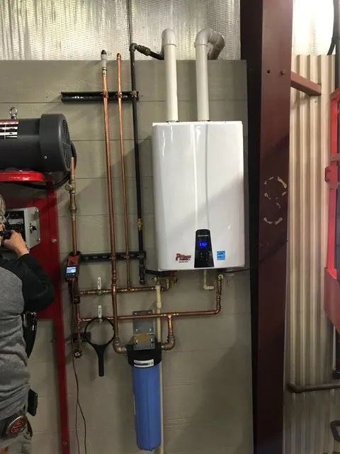 Wall-mounted white tankless water heater with copper pipes in a utility room; person partially visible on the left.