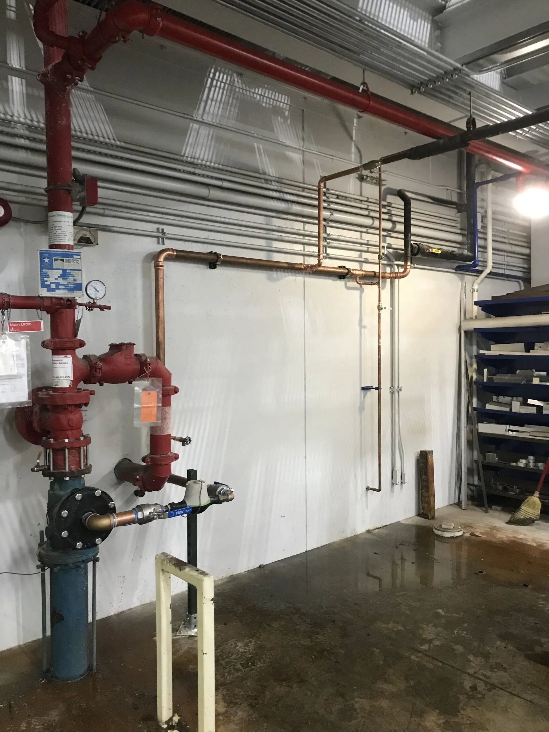 Industrial utility room with red fire pipes, white wall panels, wet concrete floor, and metal shelving