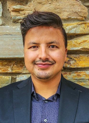 A man in a suit and blue shirt is standing in front of a stone wall.