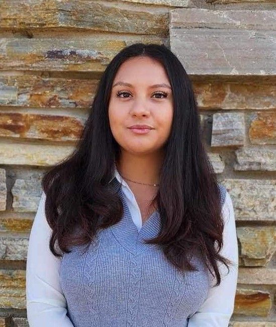 A woman in a blue vest and white shirt is standing in front of a stone wall.