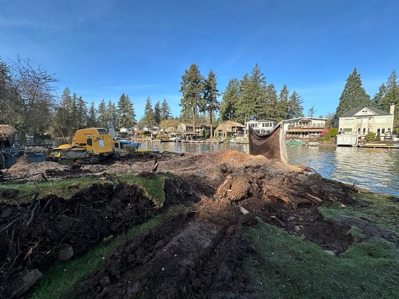 Construction site on a waterfront property with excavator; houses and lake in the background.