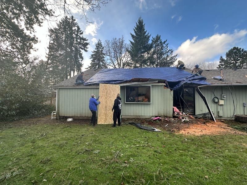 Workers repairing a damaged house roof.  Blue tarp covers some roof area. Two people hold plywood. Overcast sky.