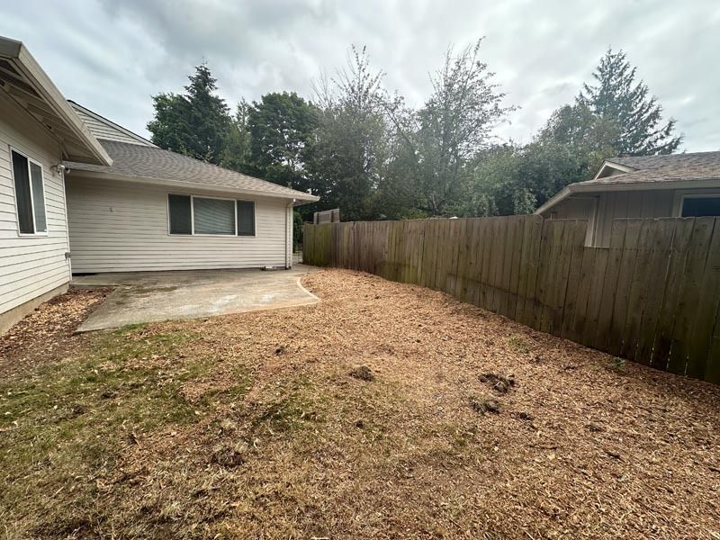 Backyard with concrete patio, wood fence, and debris on the ground; houses in the background under a cloudy sky.