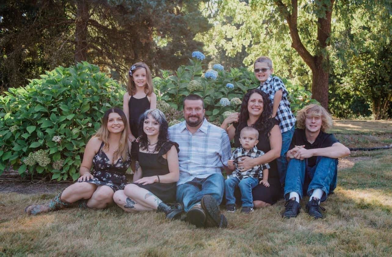 Family poses outdoors on grass, under trees and near bushes; smiles.