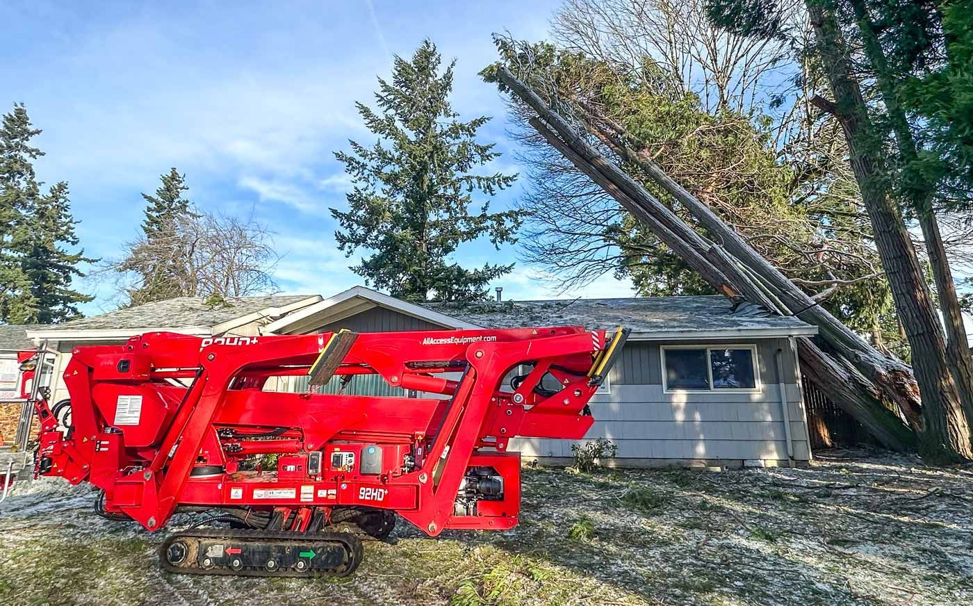 Red tree-clearing machine in front of a house, trees leaning on roof. Snowy ground, blue sky.