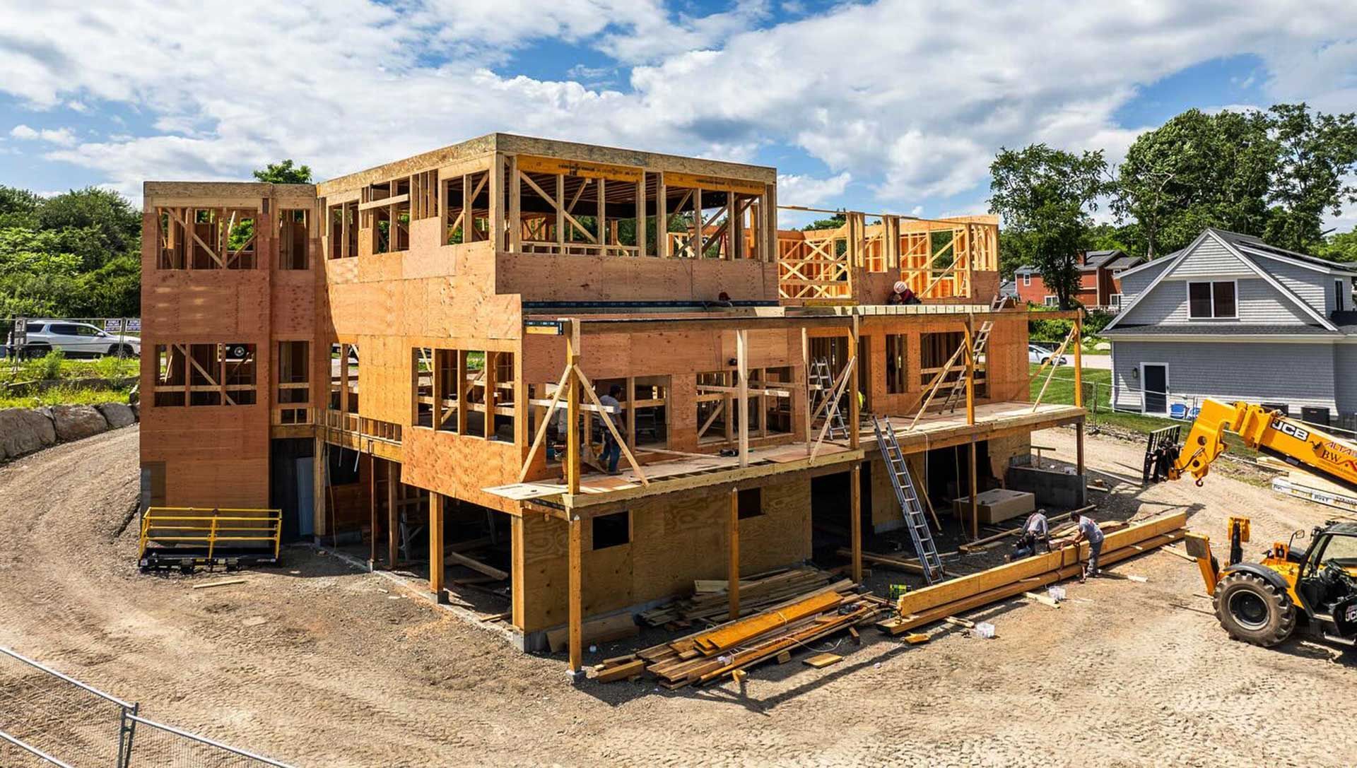 Construction of a multi-story wooden building on a dirt lot; a backhoe sits nearby.