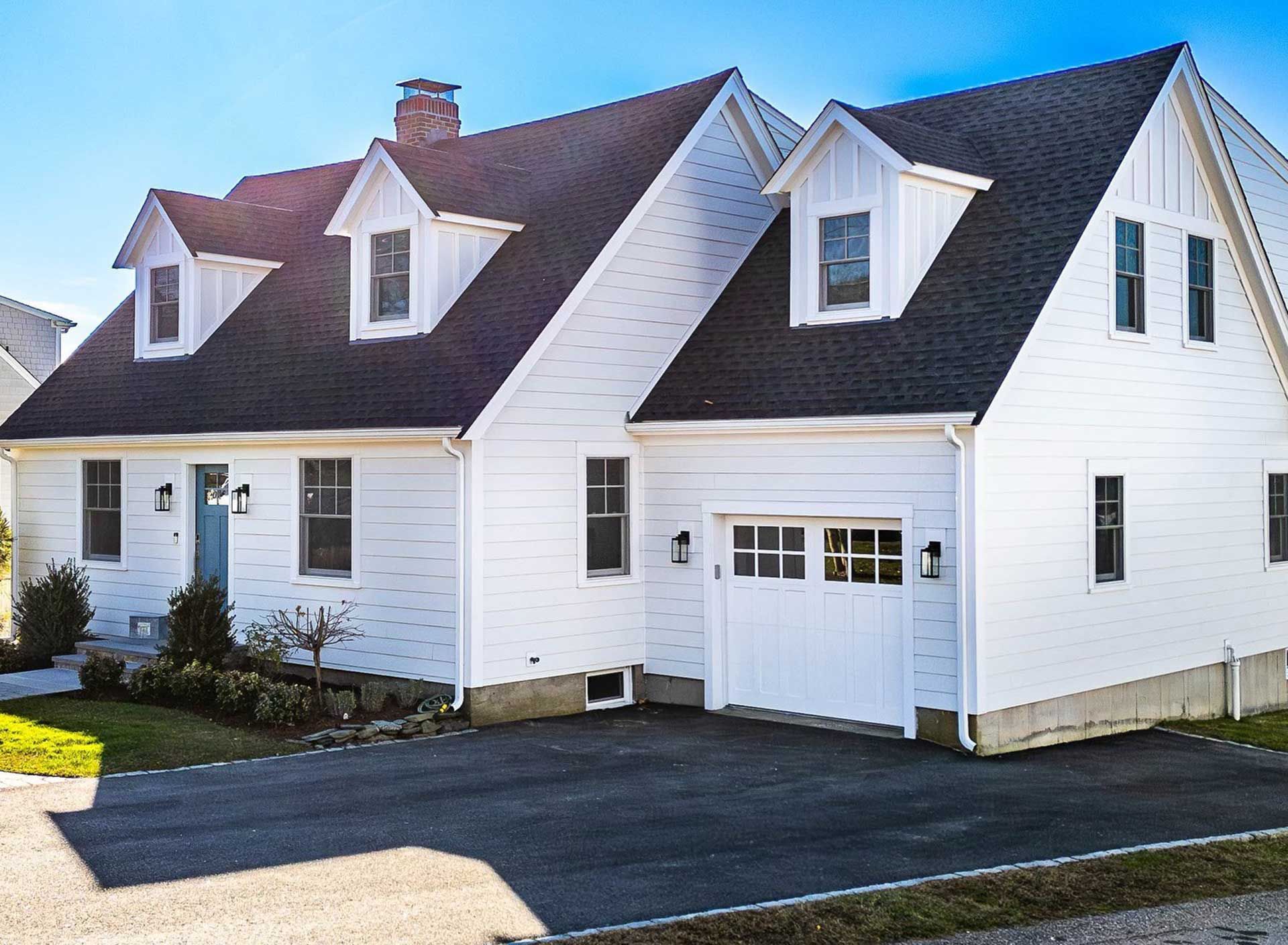 White house with black roof, three dormers, blue door, and attached garage on a sunny day.