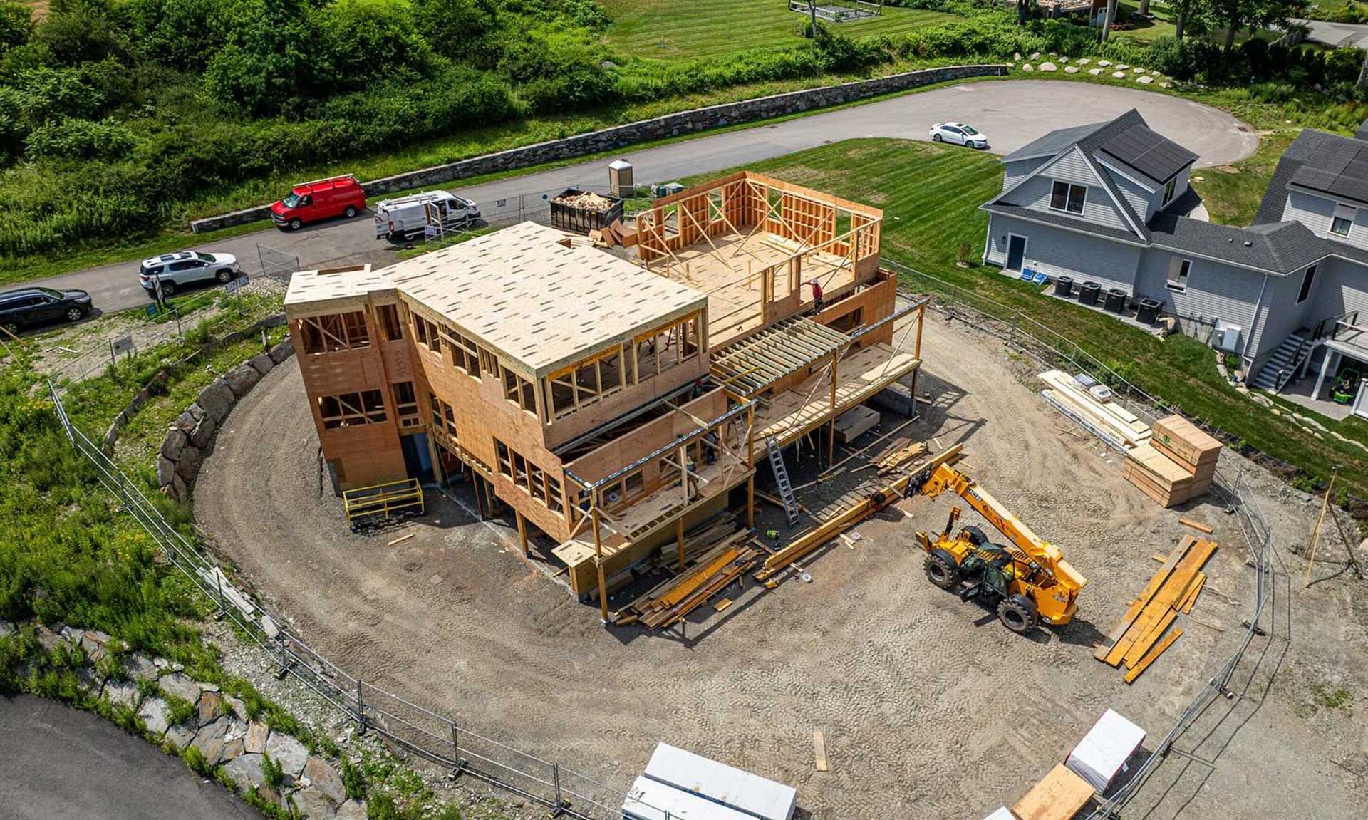 Construction of a two-story wooden framed house, with heavy machinery on a dusty lot, next to finished homes and a road.