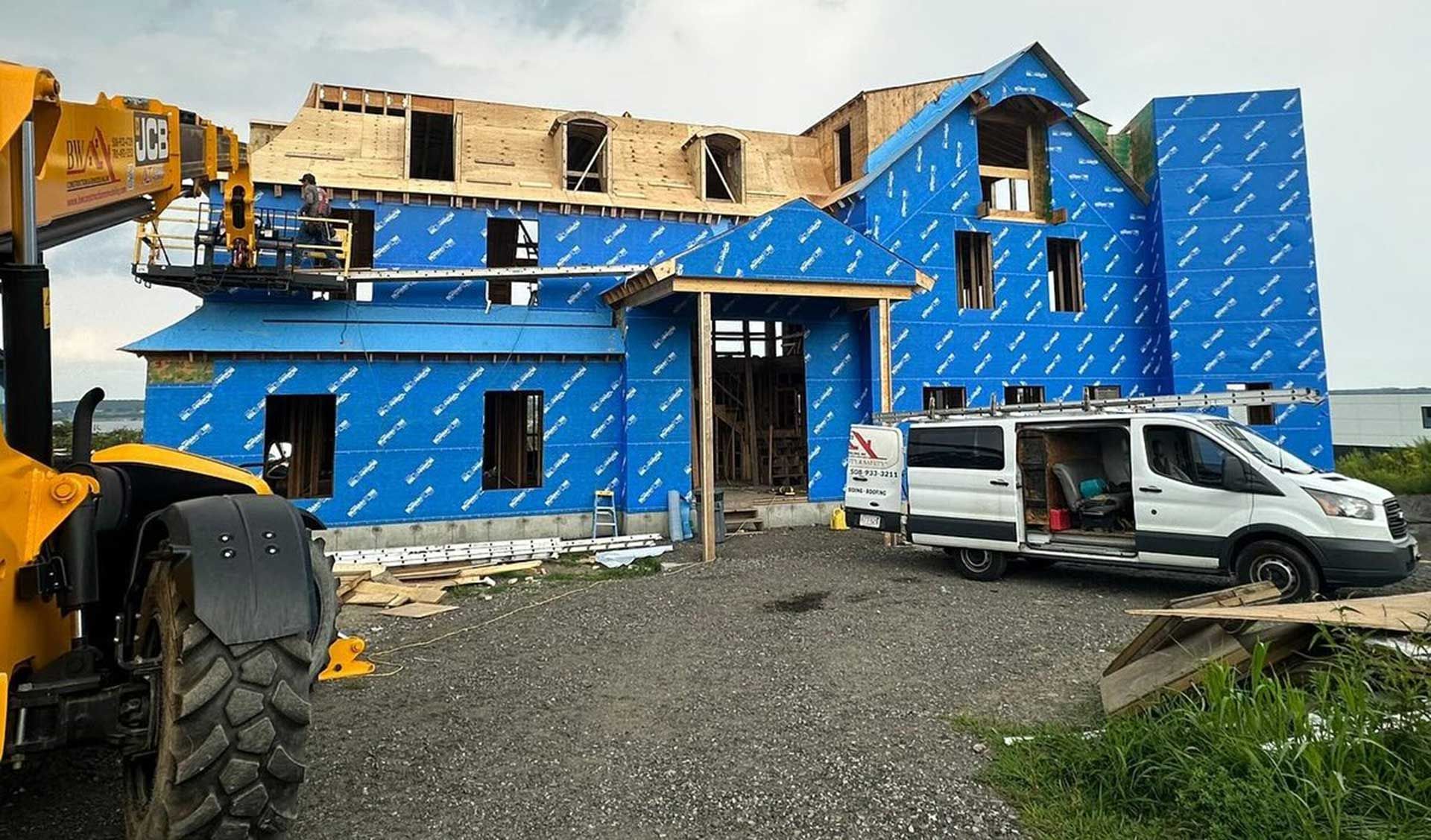 House under construction, blue weather-resistant barrier, front door frame, white van, and construction equipment.