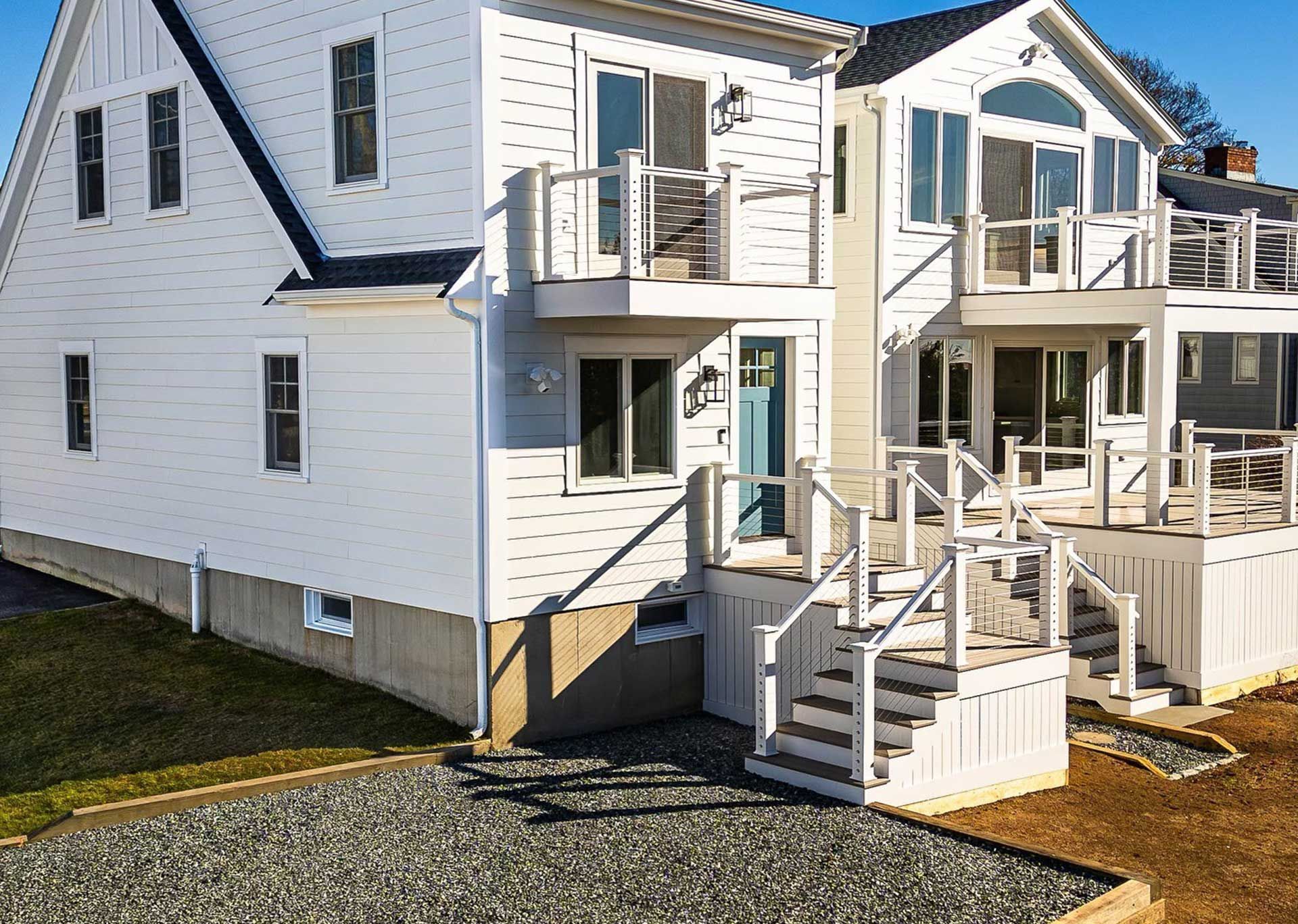 White house with decks, light blue door, gray gravel driveway, blue sky.