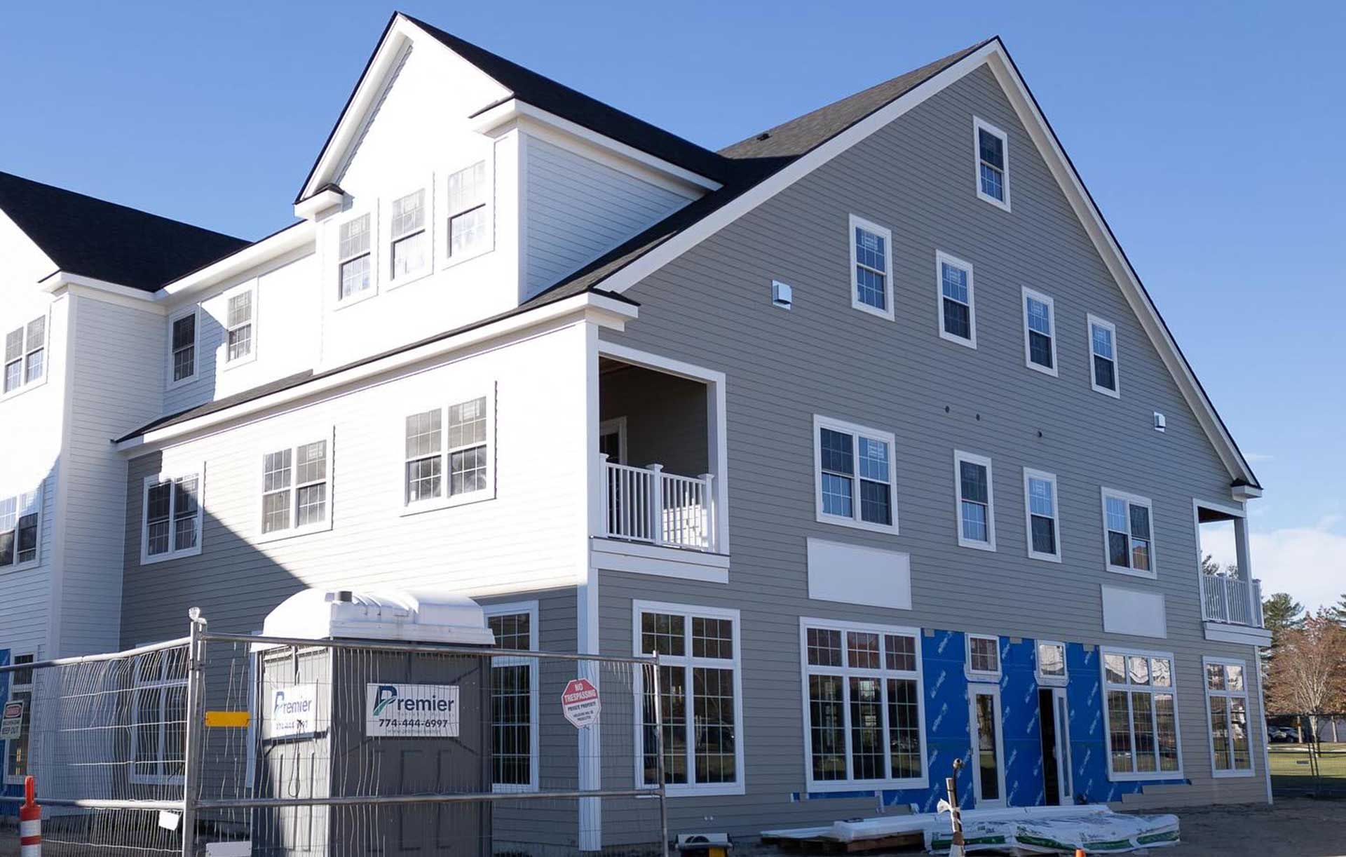 Construction site, three-story building with gray siding, large windows, and unfinished facade under a clear, sunny sky.