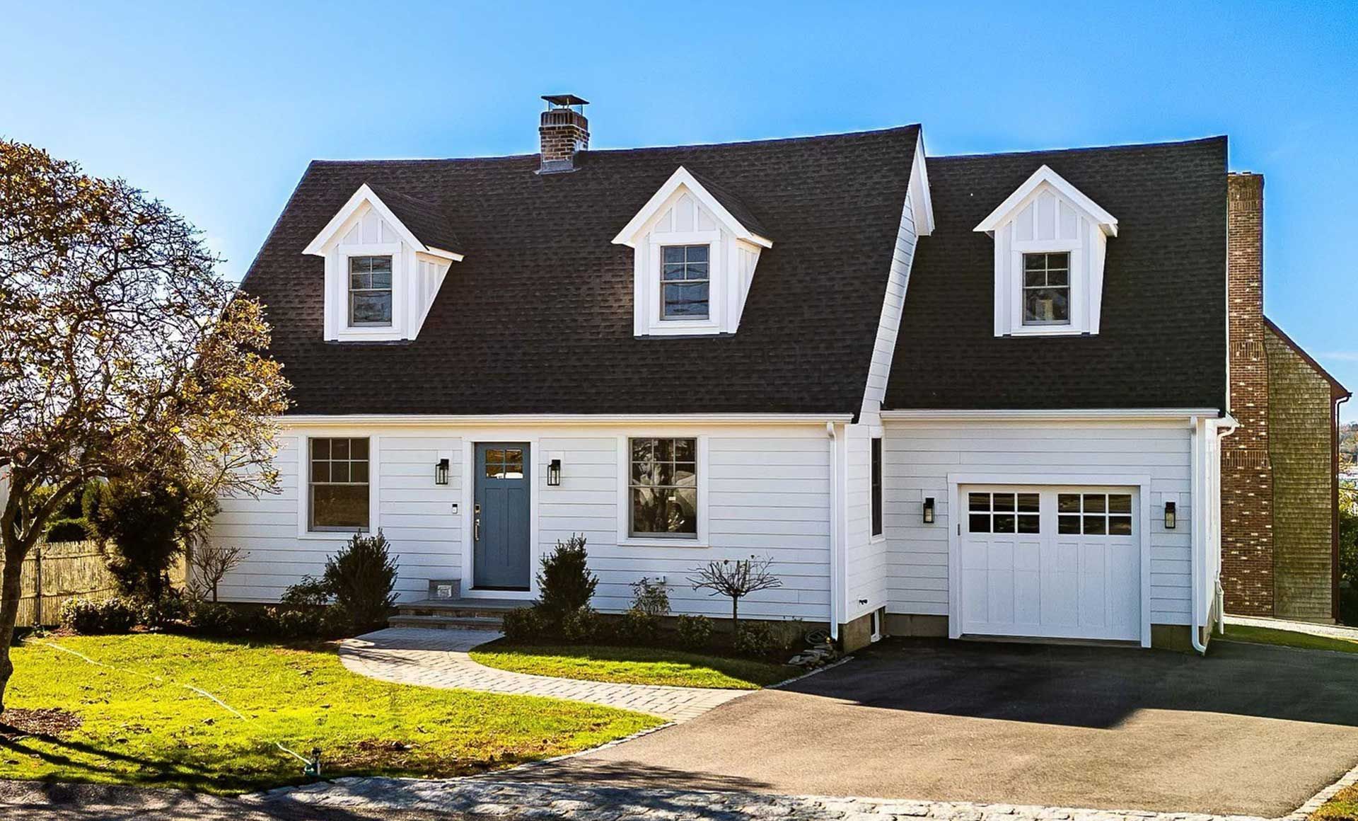 White house with a dark roof, three dormer windows, a blue door, and a garage.