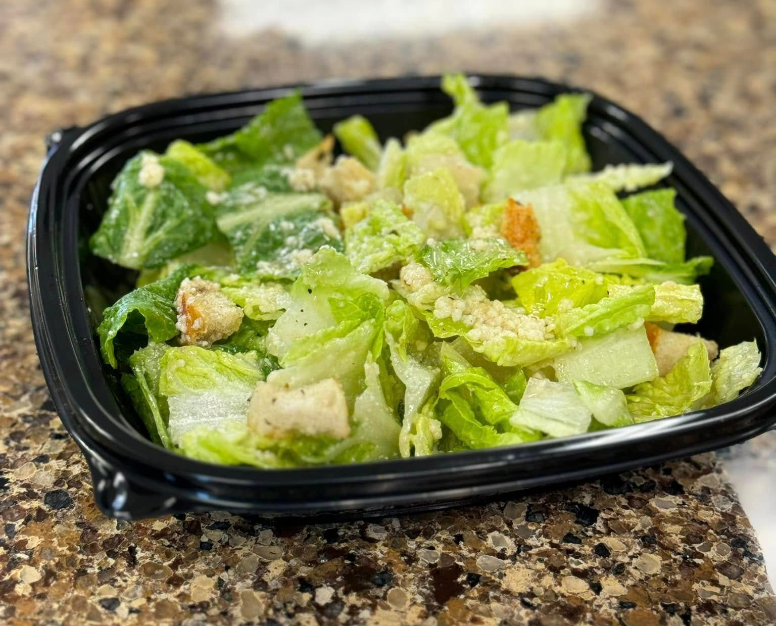 Caesar salad with lettuce, croutons, and dressing in a black takeout container on a countertop