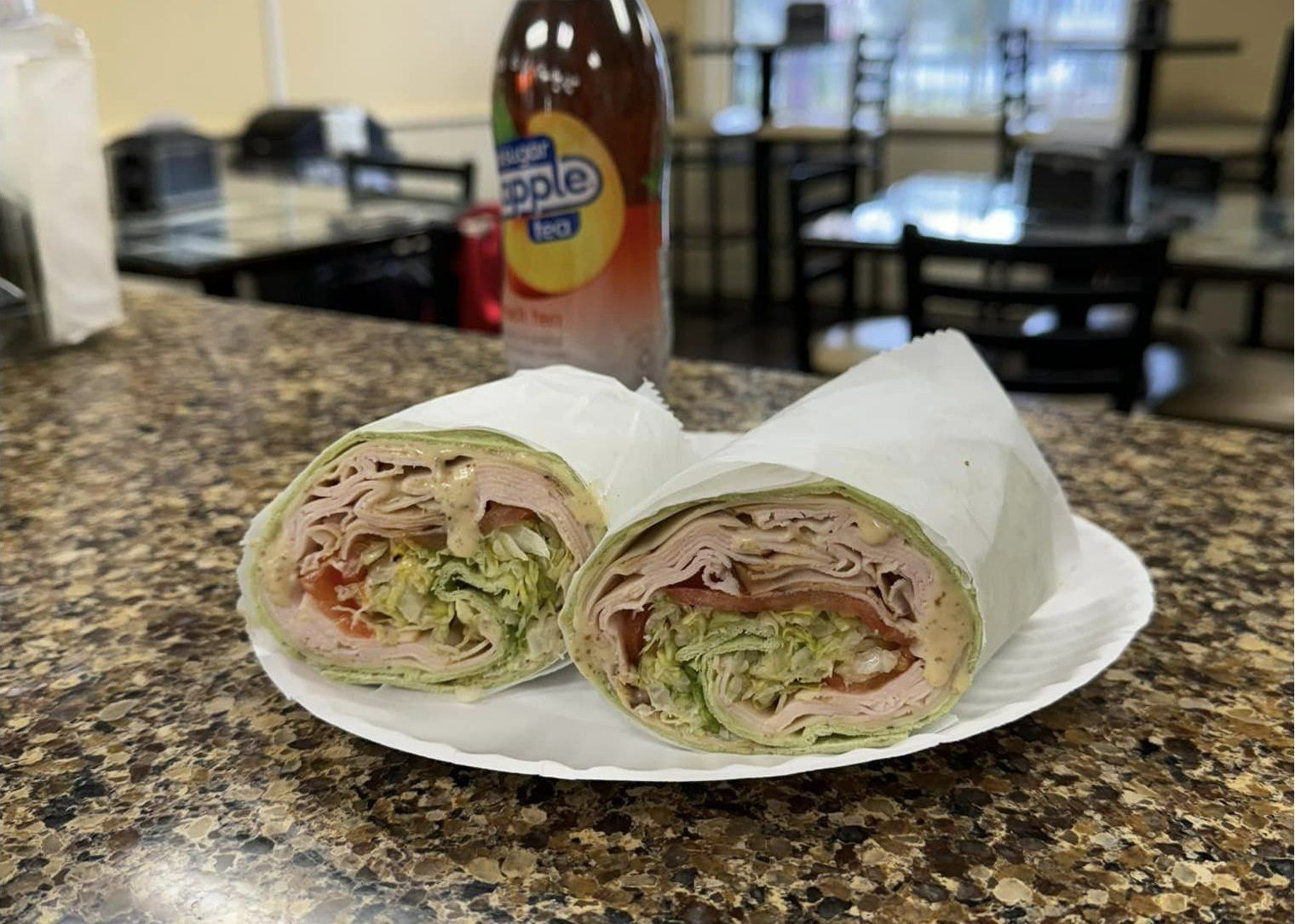 A sandwich is sitting on a white plate on a counter next to a bottle of apple juice.