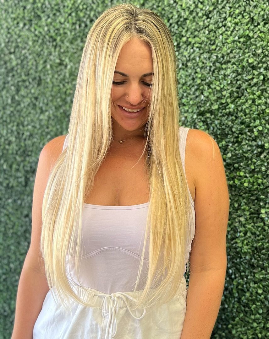 Woman with long, crimped blonde hair, smiling in front of a green wall, wearing a white top.