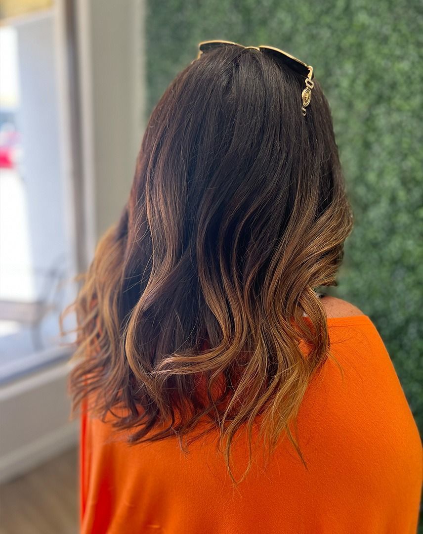 Woman with dark hair and blonde ombre, wearing an orange top, posing in front of a green wall.