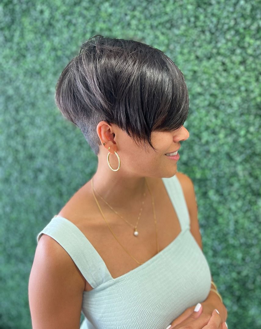 Woman with a dark, layered pixie cut, smiling. Wearing a light blue top, gold hoops, and a necklace. Set against a green backdrop.