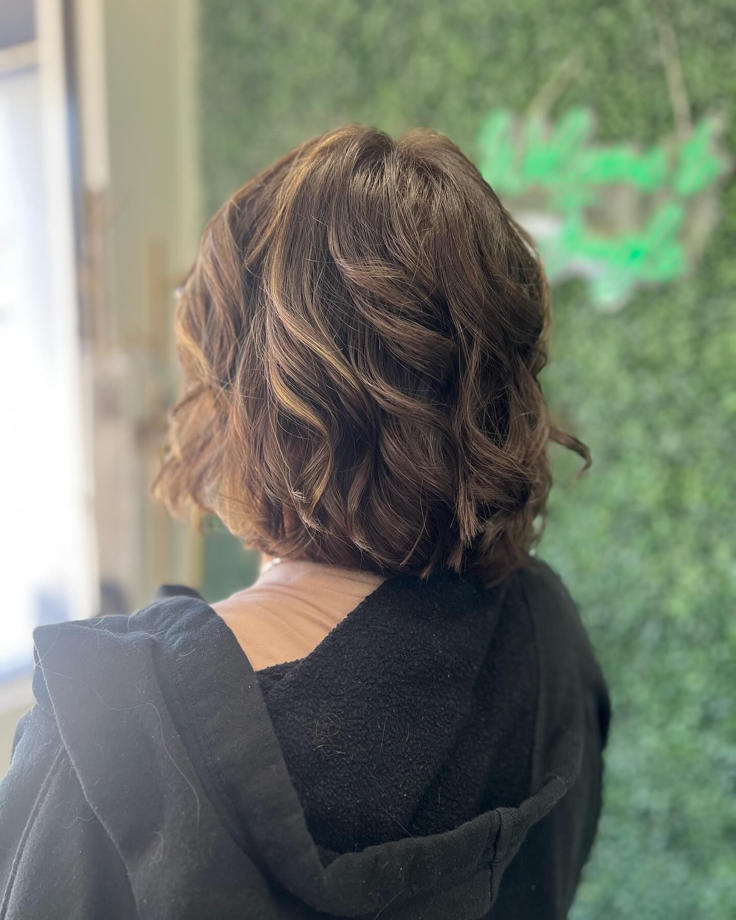 Woman with shoulder-length brown wavy hair, back view. Wearing a black top in front of a green wall.