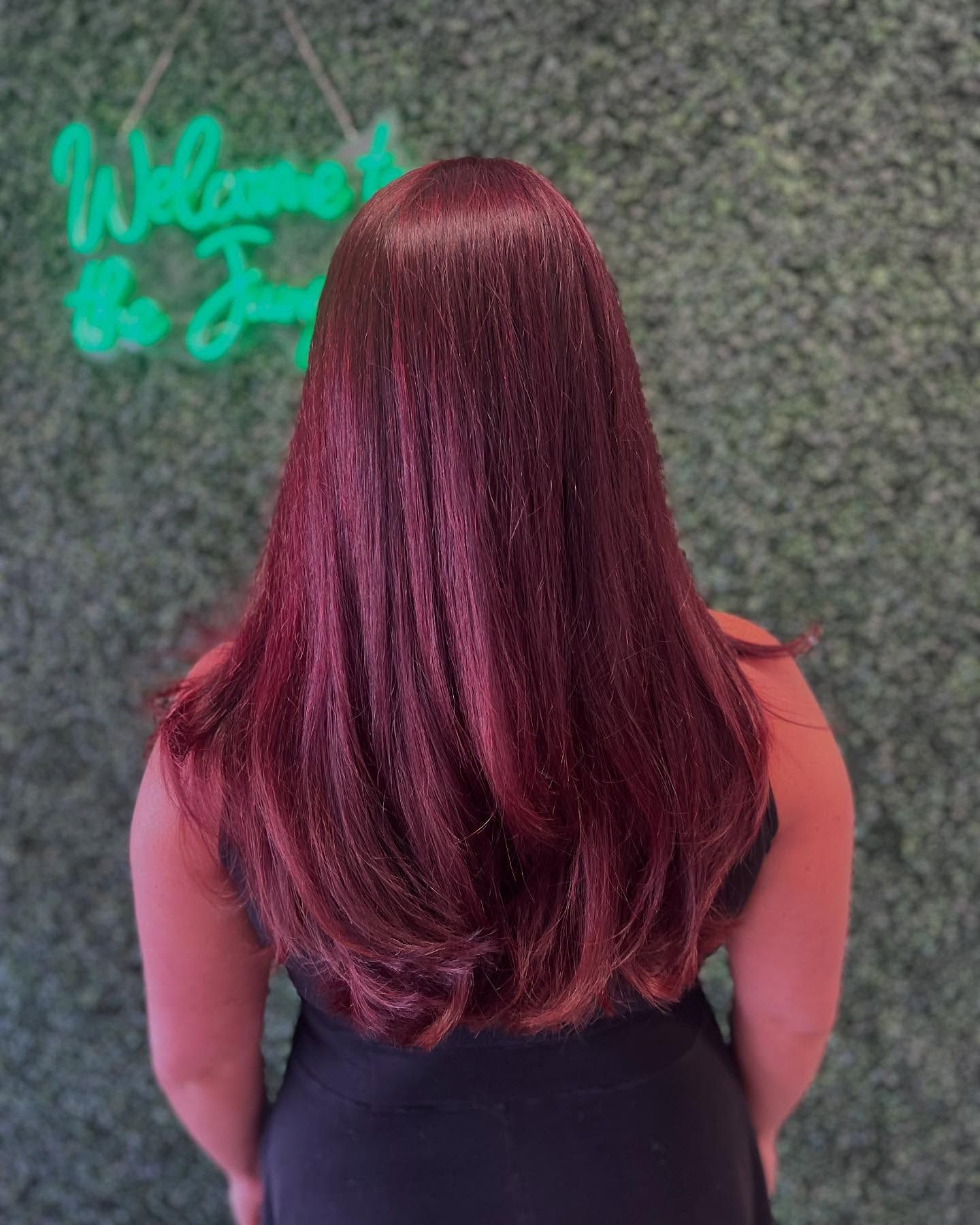 Woman with long, straight, burgundy hair, black top, standing in front of a green wall, lit neon sign.