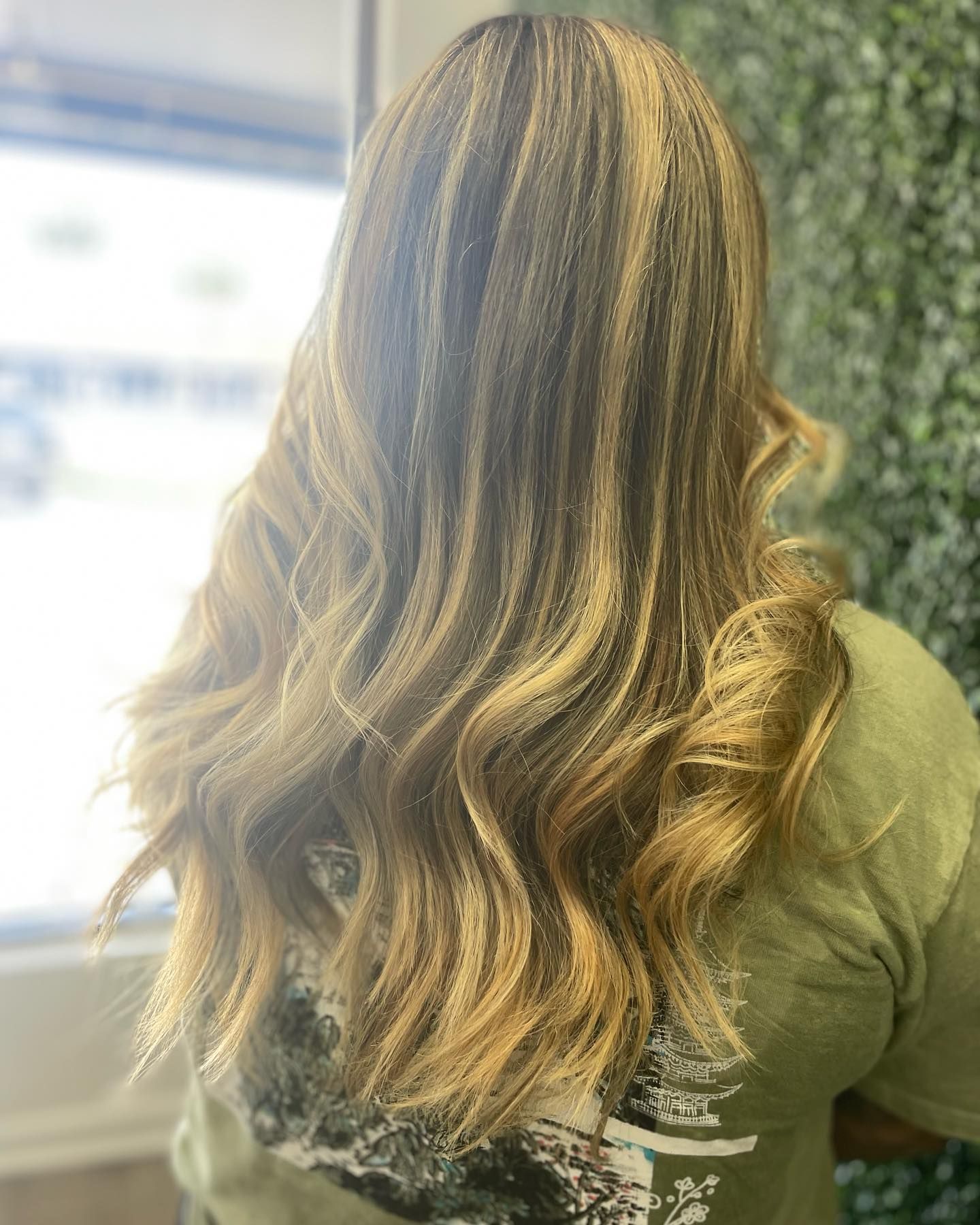 Woman with long, wavy blonde and brown highlighted hair, wearing a green shirt, standing in a salon.
