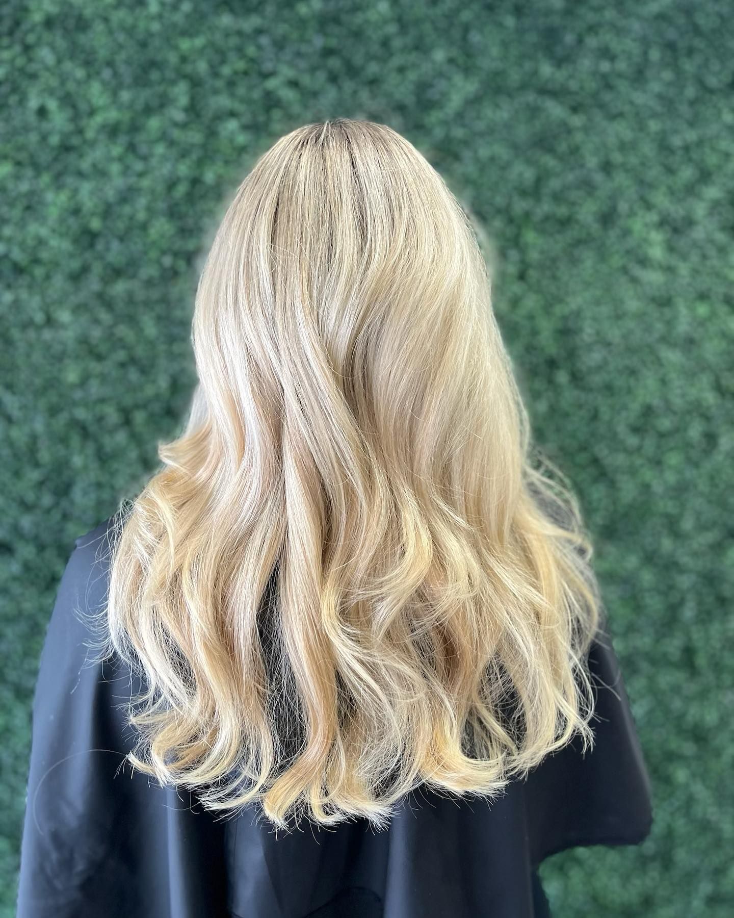 Blonde wavy hair cascading over a black cape, against a green hedge background.