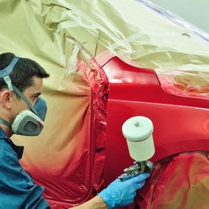 A man wearing a mask and gloves is spray painting a red car.