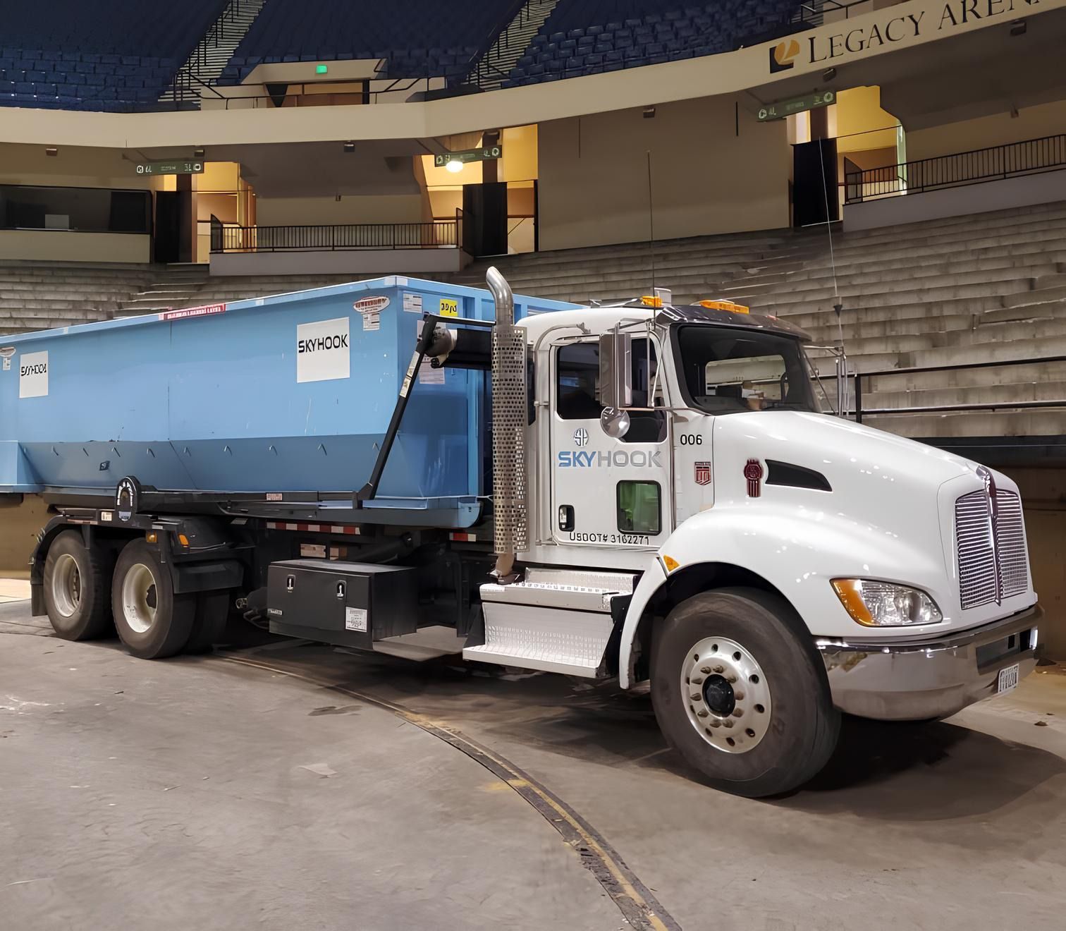 White semi-truck with a blue dumpster in an arena. The truck is parked indoors near a curved ramp.