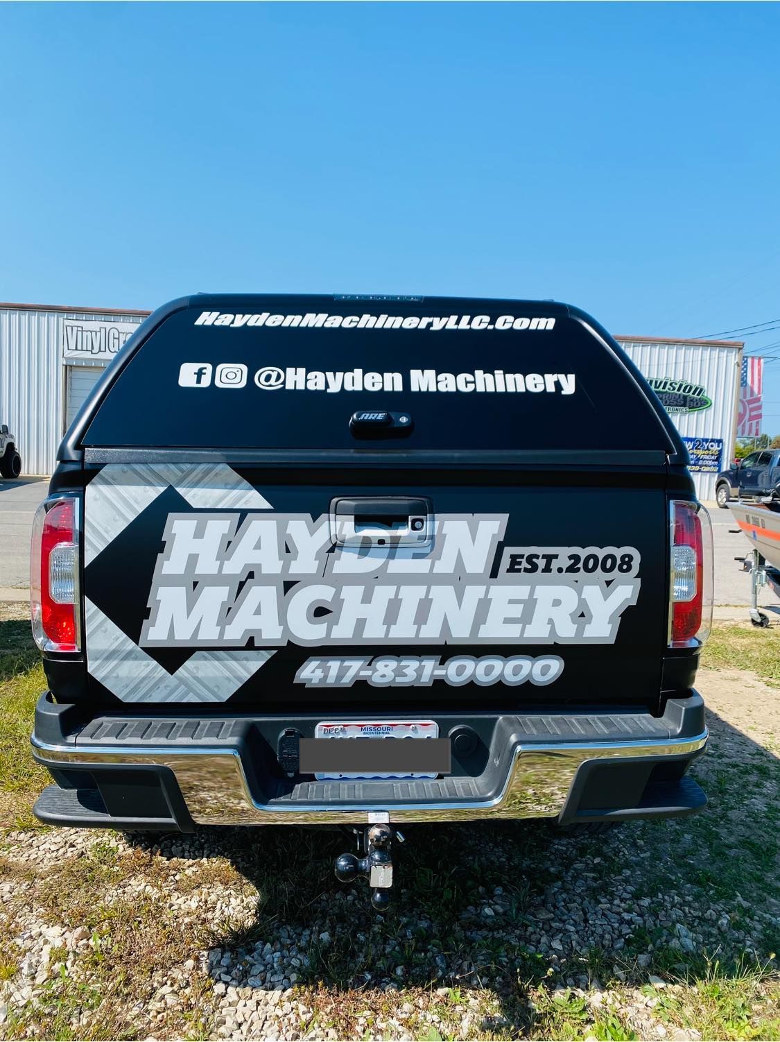 Rear of black Hayden Machinery pickup truck with white logo decals parked outdoors on gravel.