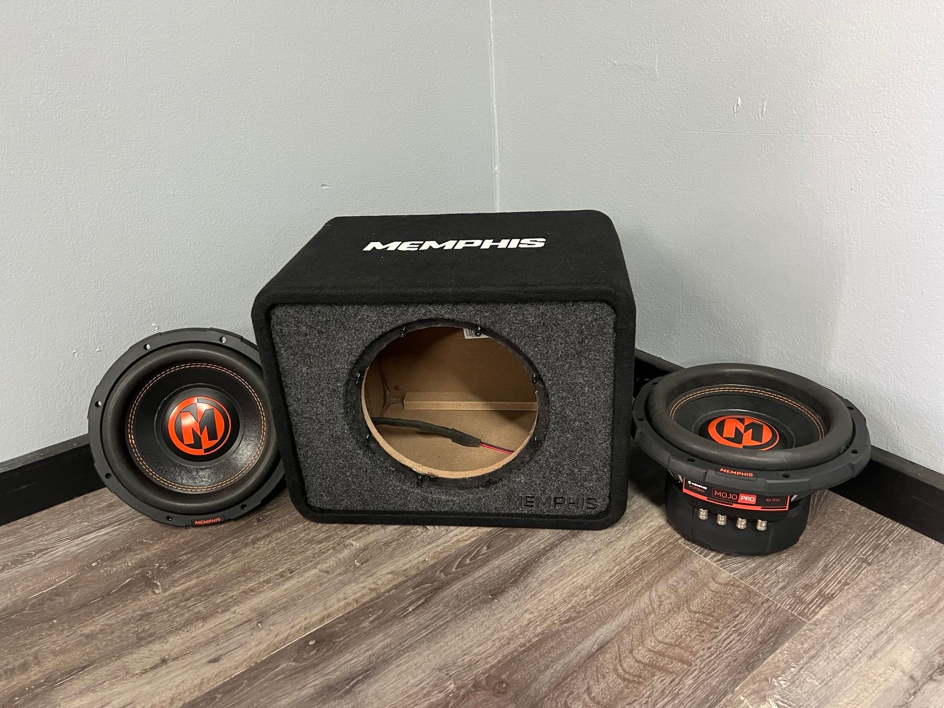 Two car speakers and a subwoofer box on a wood floor against a gray wall