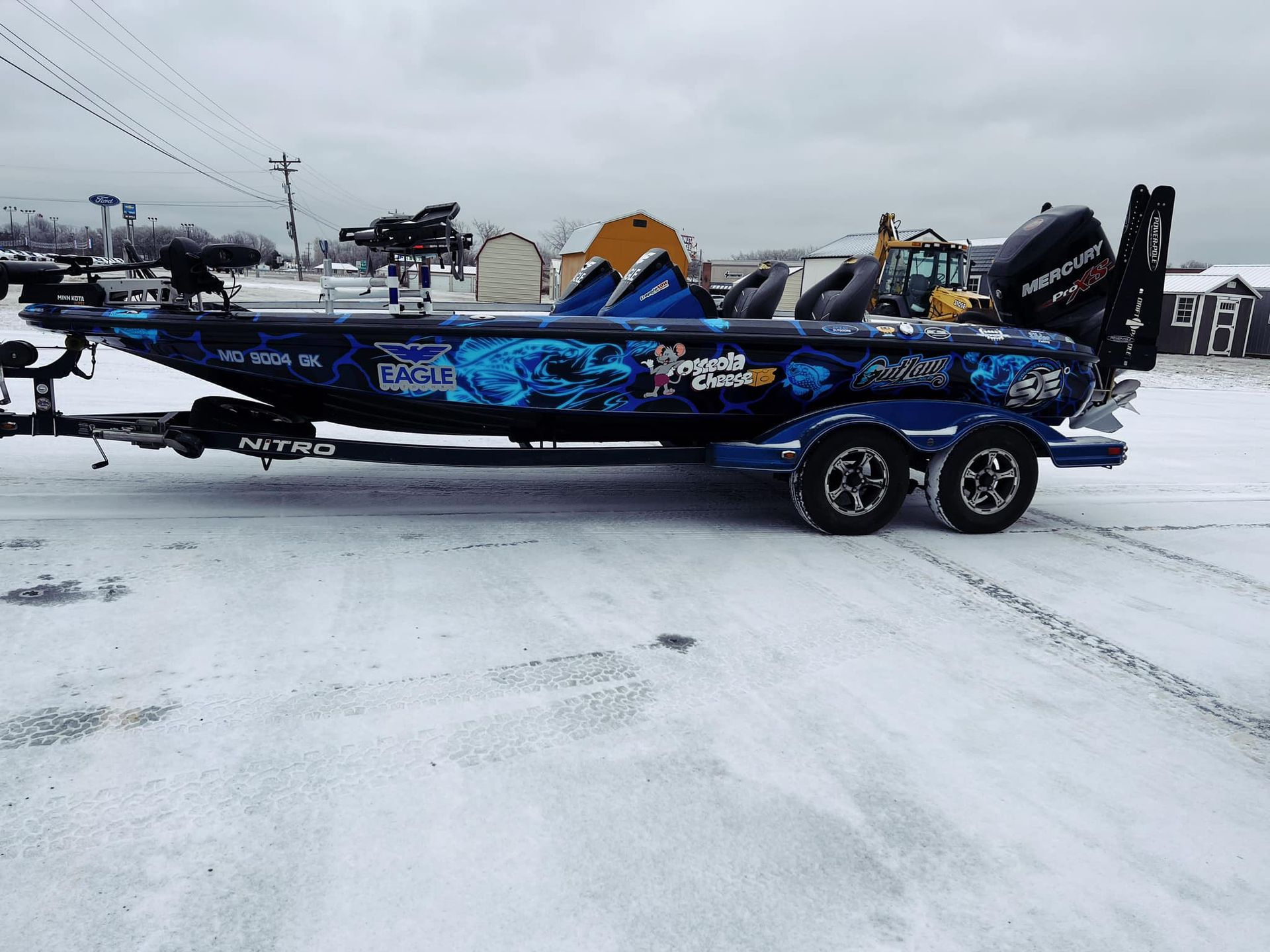 Blue bass boat on a trailer in a snowy parking lot at an outdoor event