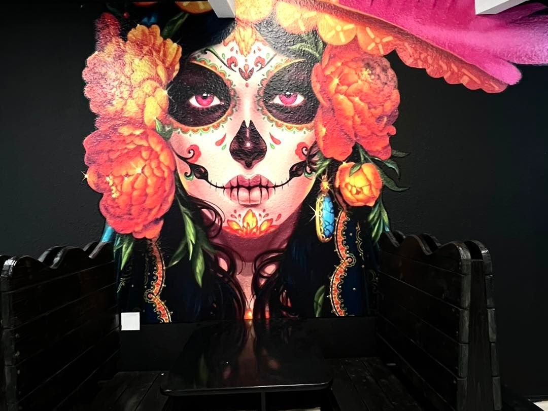 Person in elaborate sugar skull makeup with orange flowers and colorful hat against a dark background