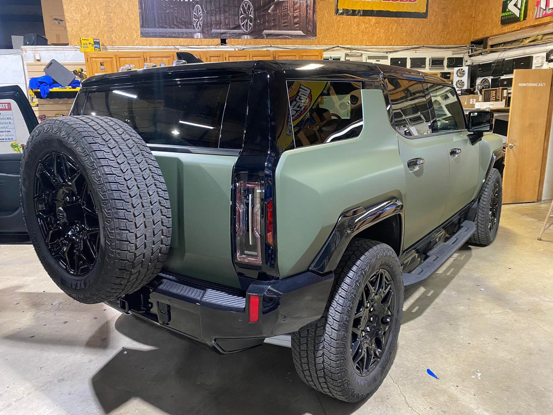 Matte green SUV in a garage, viewed from the rear-left with a spare tire mounted on the back.