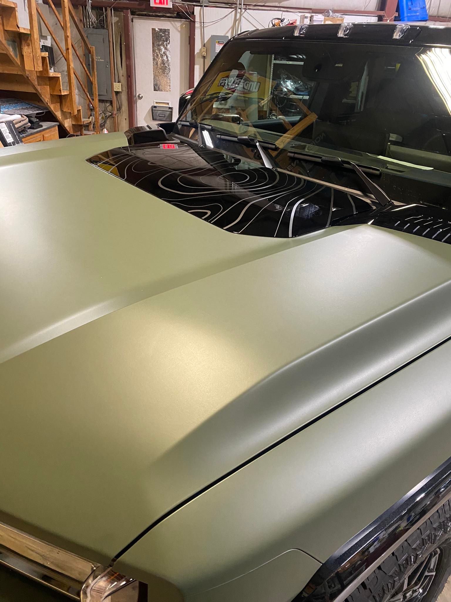 Matte olive-green SUV hood in a garage, with a black windshield and workshop shelves in the background