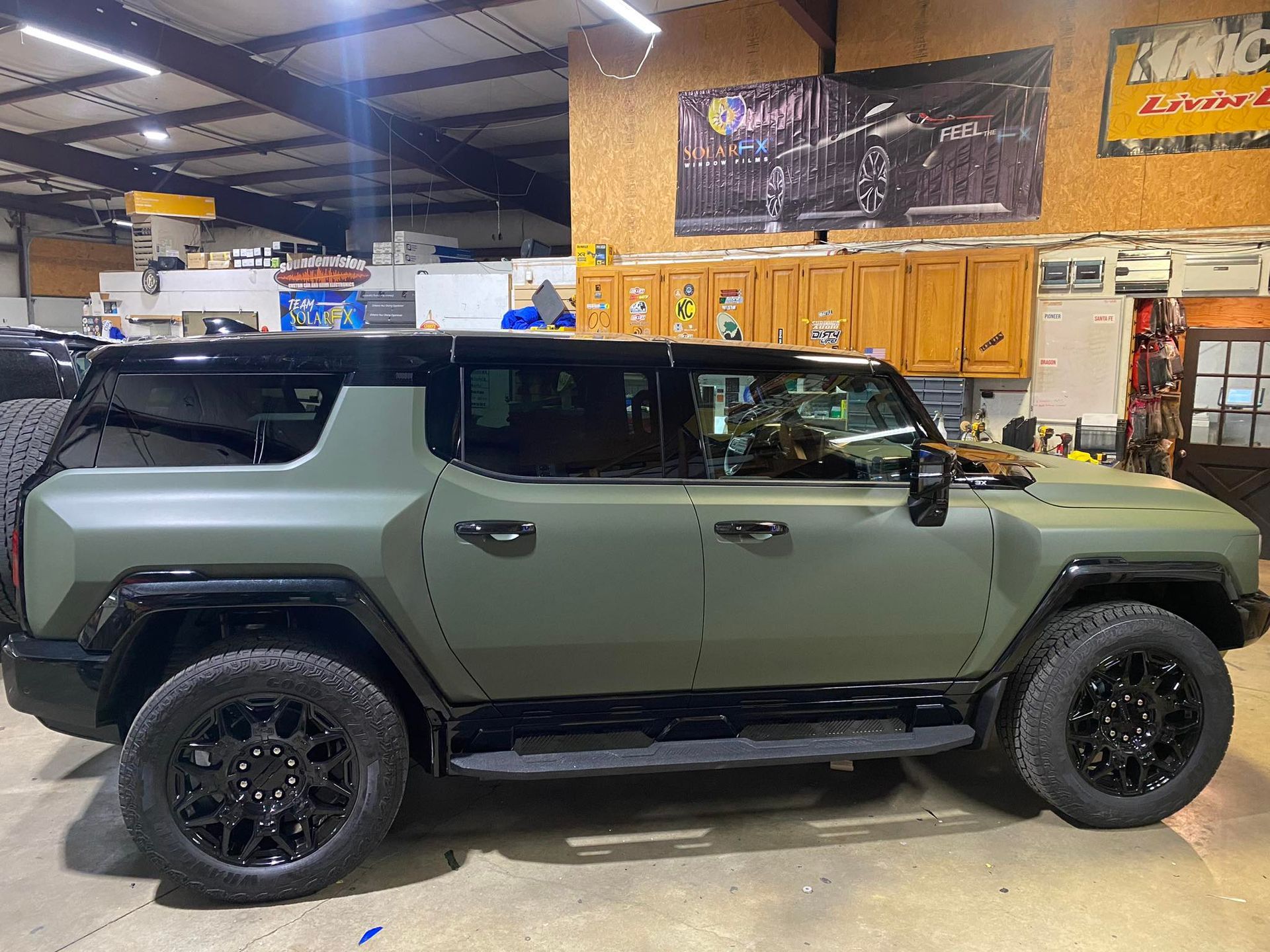 Olive green SUV in a garage with wood cabinets and a black roof rack, side view.